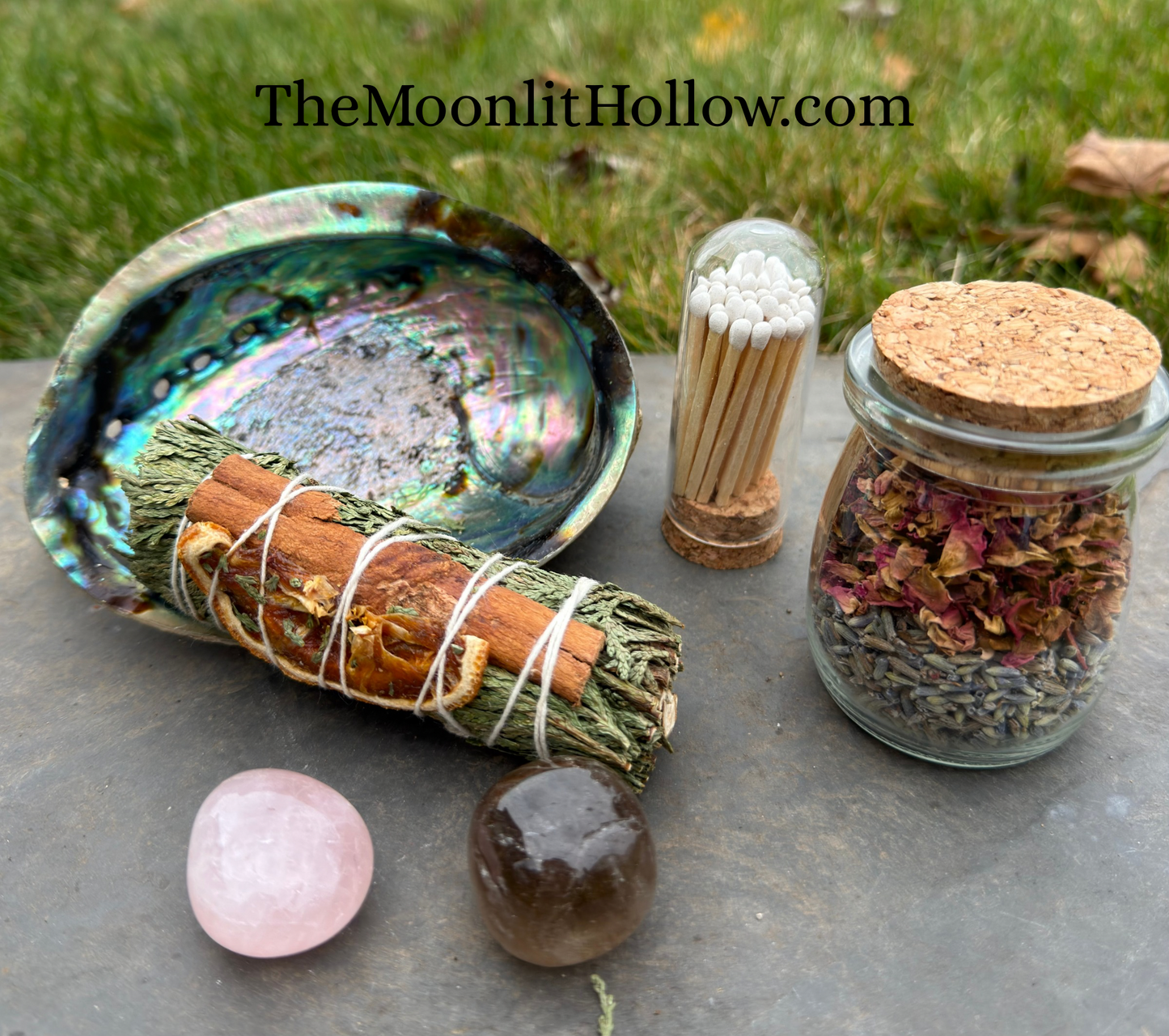 “Witchcraft home blessing kit arranged on stone: abalone shell with cedar bundle, match cloche, crystal pair, and herbal jar.”